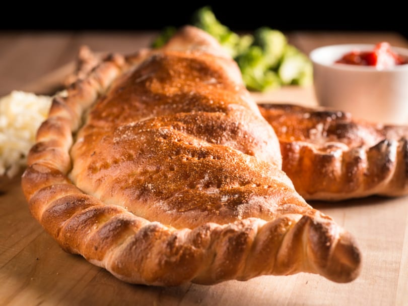 Golden-brown calzone on a wooden board with greens and a small bowl of sauce.