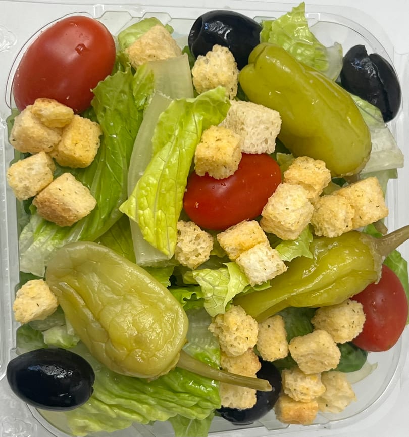 A fresh salad with lettuce, tomatoes, black olives, green peppers, and croutons in a clear plastic container.