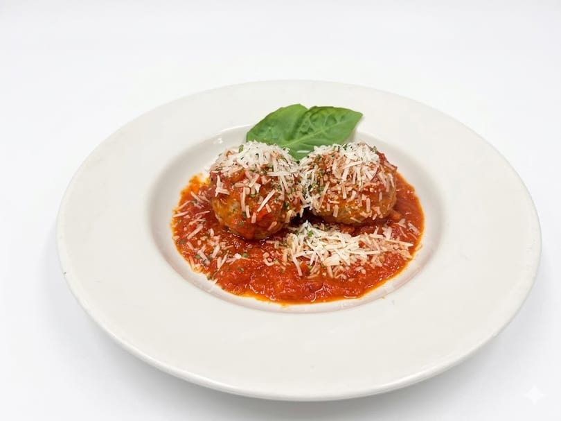 Two meatballs covered in red sauce, parmesan cheese, and parsley, with a basil leaf on a white plate.